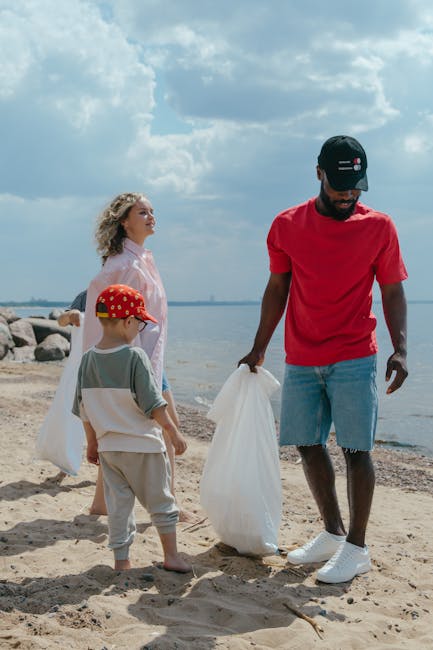 A group of three people, including an adult man and two children, are standing on a sandy beach beside a body of water under a partly cloudy sky. The man, dressed in a red T-shirt, light blue denim shorts, and white sneakers, is holding a white plastic bag and appears to be collecting litter. One child, a young boy wearing a grey T-shirt, beige pants, and a red cap with white spots, is also holding a white plastic bag and looking towards the man. The other child, a girl with curly blonde hair, is wearing a light pink jacket and white top, standing slightly behind the boy, observing the scene. Behind them, large rocks line the shoreline, with the water extending to the horizon. The image captures a moment of environmental cleanup in a natural outdoor setting, demonstrating responsible waste disposal and the importance of maintaining clean coastlines. The natural environmental context and waste collection activity suggest a focus related to rubbish removal and community efforts to manage litter in outdoor spaces, as represented by Rubbish Removal Brompton.