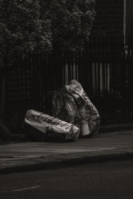 The image depicts two large black plastic garbage bags leaning against a black metal fence on a paved street or driveway. The bags are tightly sealed and appear to contain bulky waste materials, with one bag slightly crumpled and the other more stretched. Behind the fence, there are dense green bushes or trees, partially visible at the top of the image. The scene is outdoors, illuminated by natural daylight, and the setting suggests a private property or urban environment where waste is temporarily stored before collection. The overall composition emphasizes the typical visual of bulky waste ready for disposal or private rubbish collection services, aligning with the context of alternative rubbish removal and waste management options that companies like Rubbish Removal Brompton offer.