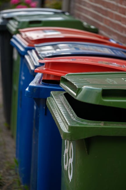 A row of five large wheelie bins lined up outdoors along a pavement, positioned against a brick wall on the right. The bins are made of plastic with textured surfaces and fitted with secure lids. The closest bin is green, with a closed lid and a white recycling symbol partially visible on its side. Behind it, there are four additional bins, each with different colored lids: red, blue, red, and dark green, all matching the color coding for waste and recycling collection. The bins are arranged in a neat line, extending into the distance, with the lids all closed, indicating they are ready for collection. The environment appears to be a residential or communal area, with natural daylight providing even lighting, highlighting the textures of the plastic and the surrounding brickwork. This scene illustrates typical arrangements for private waste handling, often associated with rubbish removal services such as those provided by Rubbish Removal Brompton, especially for alternative collection arrangements outside of local authority services.