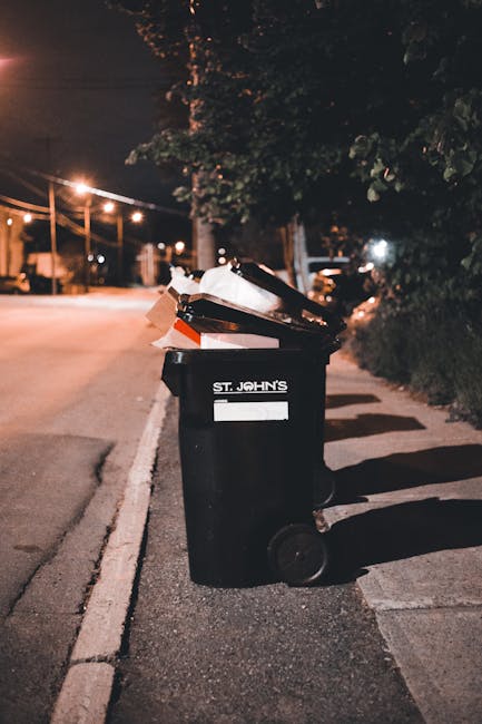 The image depicts two large black plastic garbage bags leaning against a black metal fence on a paved street or driveway. The bags are tightly sealed and appear to contain bulky waste materials, with one bag slightly crumpled and the other more stretched. Behind the fence, there are dense green bushes or trees, partially visible at the top of the image. The scene is outdoors, illuminated by natural daylight, and the setting suggests a private property or urban environment where waste is temporarily stored before collection. The overall composition emphasizes the typical visual of bulky waste ready for disposal or private rubbish collection services, aligning with the context of alternative rubbish removal and waste management options that companies like Rubbish Removal Brompton offer.