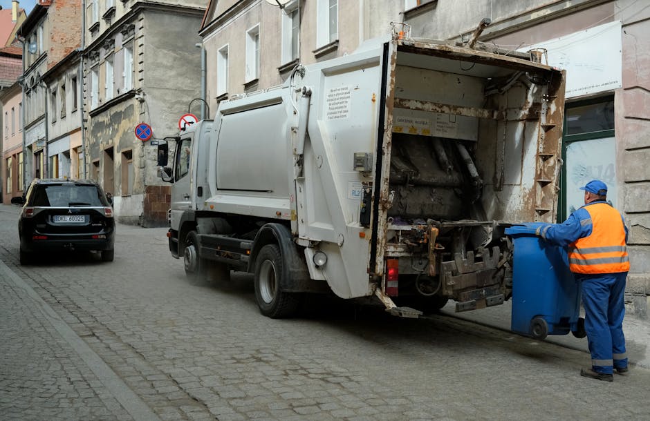 A street scene shows a white waste collection vehicle actively engaged in rubbish removal, with a large open rear compactor section partially rusted and visibly used for collecting waste. The vehicle is parked close to a row of old, weathered buildings with a mixture of brick and plaster facades, some showing signs of aging and peeling paint. An employee dressed in high-visibility orange and blue uniform, wearing a blue cap, is in the process of emptying a medium-sized blue recycling or waste bin into the back of the vehicle, with their hands gripping the bin's handles. To the left of the truck, a dark grey car is parked on the cobbled street, which is lined with a paving pattern typical of urban residential areas. The scene is outdoors, with natural light illuminating the street, and the overall environment reflects a typical urban rubbish collection operation, indicating an on-site waste disposal or private rubbish removal service. The setting underscores local, independent waste management, consistent with services offered by Rubbish Removal Brompton.