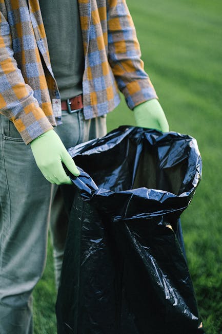 A person wearing a casual outfit that includes a checkered shirt with shades of yellow, blue, and gray, over a dark T-shirt, and green rubber gloves, is standing outdoors on a grassy area. They are holding open a large black rubbish bag made of durable plastic, with the bag's top turned outward, displaying its empty interior. The scene suggests a waste collection or rubbish removal activity, likely related to garden or outdoor cleaning. The background features lush, green grass, indicating a well-maintained outdoor environment. The image captures the process of preparing a rubbish bag for collecting waste, consistent with waste management or rubbish removal services. The scene is well-lit with natural daylight, emphasizing the textures of the plastic bag, the gloves, and the person's clothing, all aligned with the visual context of private waste handling and rubbish collection activities.