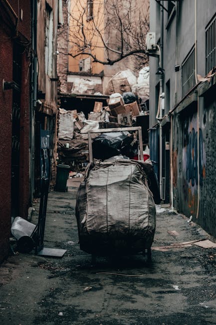 A street scene shows a white waste collection vehicle actively engaged in rubbish removal, with a large open rear compactor section partially rusted and visibly used for collecting waste. The vehicle is parked close to a row of old, weathered buildings with a mixture of brick and plaster facades, some showing signs of aging and peeling paint. An employee dressed in high-visibility orange and blue uniform, wearing a blue cap, is in the process of emptying a medium-sized blue recycling or waste bin into the back of the vehicle, with their hands gripping the bin's handles. To the left of the truck, a dark grey car is parked on the cobbled street, which is lined with a paving pattern typical of urban residential areas. The scene is outdoors, with natural light illuminating the street, and the overall environment reflects a typical urban rubbish collection operation, indicating an on-site waste disposal or private rubbish removal service. The setting underscores local, independent waste management, consistent with services offered by Rubbish Removal Brompton.