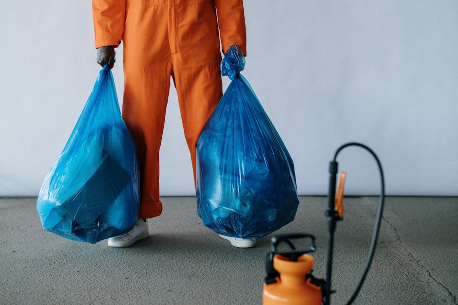 The image shows a person dressed in an orange waste collection uniform standing on a grey concrete floor, holding two large, semi-transparent blue plastic rubbish bags filled with waste materials. The person's hands are gloved, and they are wearing white footwear. In the foreground, there is a partially visible orange waste sprayer or fogging device connected to a black hose, suggesting cleaning or sanitizing activity. The background is plain and light-colored, providing a neutral setting typical of a waste management or industrial environment. This scene reflects the process of rubbish collection or disposal, which relates to services offered by companies like Rubbish Removal Brompton, specialising in independent waste removal solutions such as on-site clearance or private disposal methods.