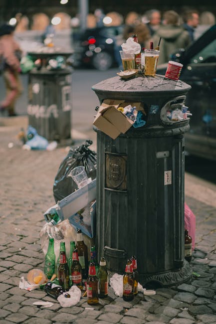 A large, dark green public litter bin with a rounded, flat top situated on a cobblestone sidewalk. The top of the bin is cluttered with various discarded items including paper coffee cups with plastic lids, an empty beer glass, a crumpled cardboard food container, and a small red container. Surrounding the base of the bin are several empty glass bottles, some spilling onto the cobbles, along with crumpled paper and plastic waste. In the background, blurred figures of pedestrians walk along the street, and a dark vehicle is partially visible on the right. To the left of the bin, there is a grey trash bag leaning against it, and a grey waste container further back in the distance. The scene suggests an environment where improper disposal of waste occurs, highlighting the importance of effective rubbish collection and disposal services such as those offered by Rubbish Removal Brompton, which are vital for maintaining cleanliness and environmental standards in busy urban areas.