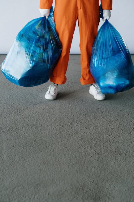 The image shows a person dressed in an orange waste collection uniform standing on a grey concrete floor, holding two large, semi-transparent blue plastic rubbish bags filled with waste materials. The person's hands are gloved, and they are wearing white footwear. In the foreground, there is a partially visible orange waste sprayer or fogging device connected to a black hose, suggesting cleaning or sanitizing activity. The background is plain and light-colored, providing a neutral setting typical of a waste management or industrial environment. This scene reflects the process of rubbish collection or disposal, which relates to services offered by companies like Rubbish Removal Brompton, specialising in independent waste removal solutions such as on-site clearance or private disposal methods.